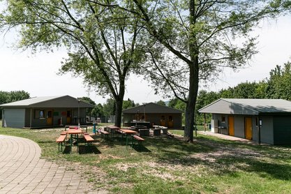 Drei Holz-Wohnhütten mit Bierbänken und Tischen davor unter zwei Bäumen 